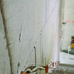 Paweł Gutowski GPJ Energia - Surowa ściana z betonu komórkowego z pomarańczowymi puszkami instalacyjnymi i wystającymi przewodami elektrycznymi w kolorach niebieskim, brązowym i zielono-żółtym, widok z dołu.