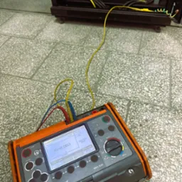 Miernik Sonel MPI-525 z podłączonymi przewodami pomiarowymi, wyświetlający wynik pomiaru rezystancji uziemienia 0,260Ω, ustawiony na posadzce z płytek, obok szafy rack z widocznym przewodem...