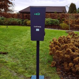 Paweł Gutowski GPJ Energia - Stacja ładowania samochodów elektrycznych CenEnergy na tle zielonego trawnika i krzewów, z widocznym budynkiem w tle.
