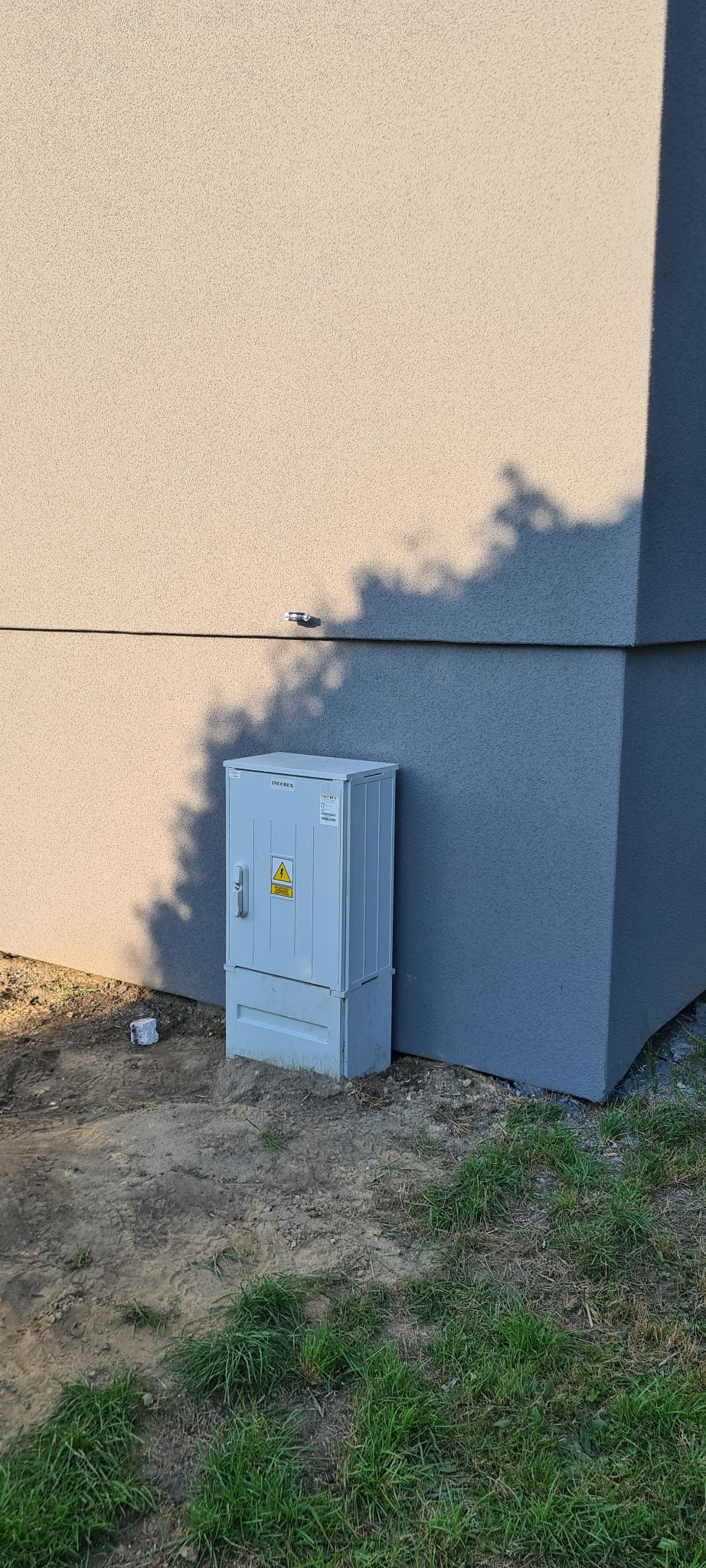 Szara skrzynka elektryczna z ostrzeżeniem o wysokim napięciu, zamontowana przy ścianie budynku z widocznym cieniem drzewa.