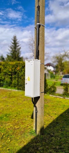 Biała skrzynka elektryczna zamontowana na betonowym słupie, z widocznymi przewodami i żółtym znakiem ostrzegawczym, na tle zielonego trawnika i drzew.
