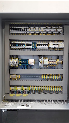 Rozdzielnica elektryczna w szarej obudowie, widoczne zabezpieczenia, styczniki, listwy zaciskowe i okablowanie, ułożone w rzędach, z widocznymi etykietami na elementach.