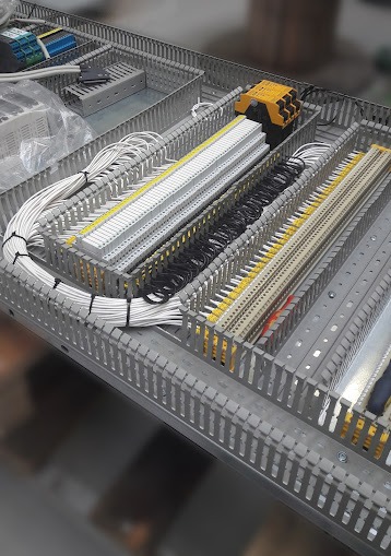 Szafa sterownicza z uporządkowanymi przewodami: białe, czarne i żółte kable podłączone do listew zaciskowych, widoczne elementy automatyki i osprzęt w metalowej obudowie z perforowanymi ściankami.