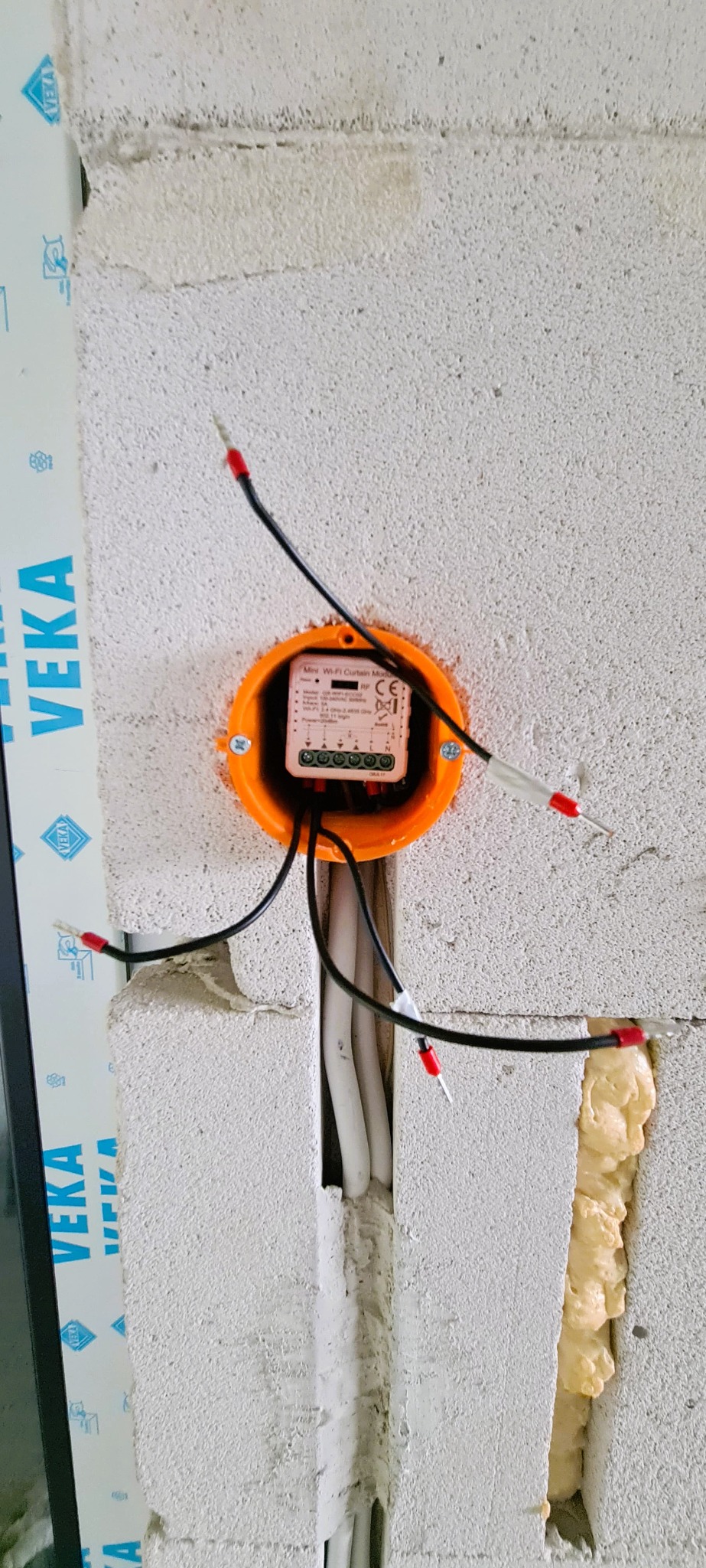 Pomarańczowa puszka instalacyjna z modułem WiFi w surowej ścianie z betonu komórkowego, widoczne przewody z końcówkami tulejkowymi oraz pianka montażowa w szczelinie obok.