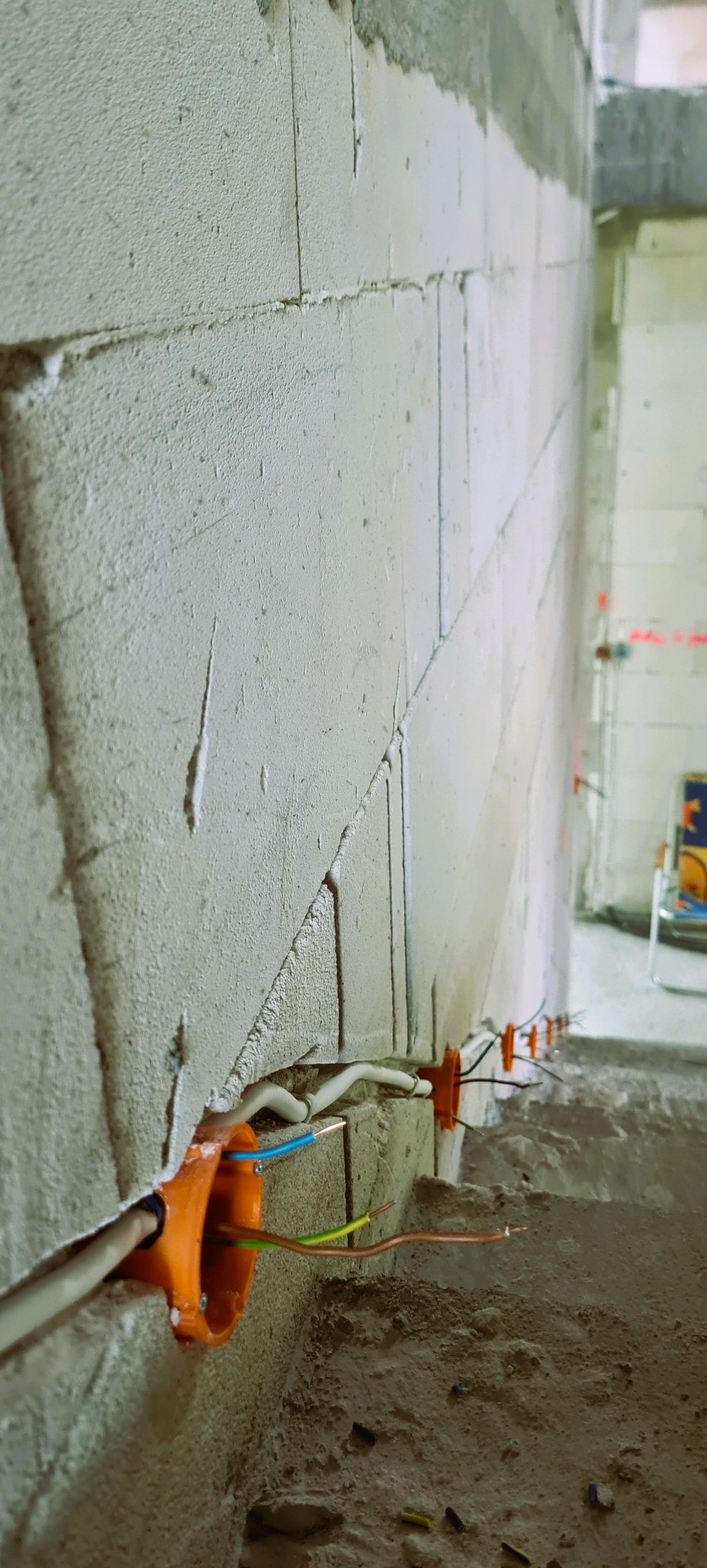 Surowa ściana z betonu komórkowego z pomarańczowymi puszkami instalacyjnymi i wystającymi przewodami elektrycznymi w kolorach niebieskim, brązowym i zielono-żółtym, widok z dołu.