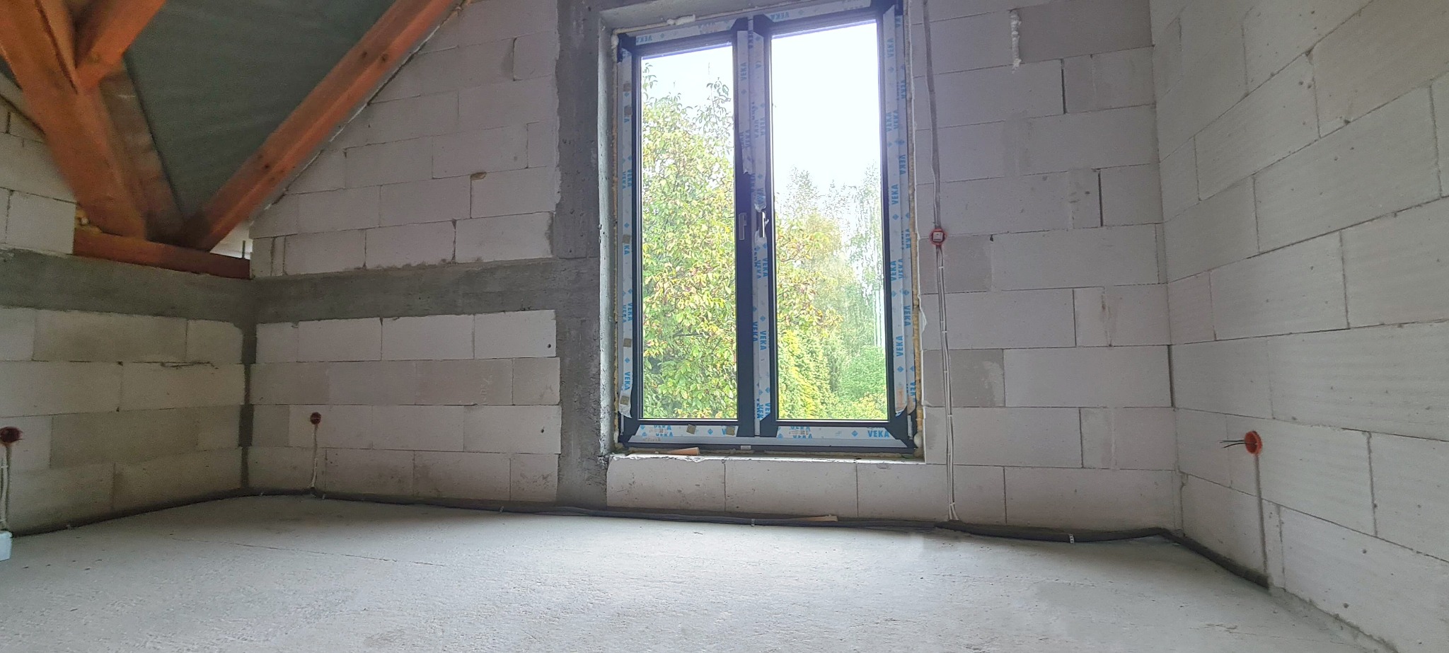 Surowy stan poddasza z oknem PVC, widoczne kable elektryczne wystające ze ściany z betonu komórkowego, drewniana konstrukcja dachu i wylewka na podłodze.