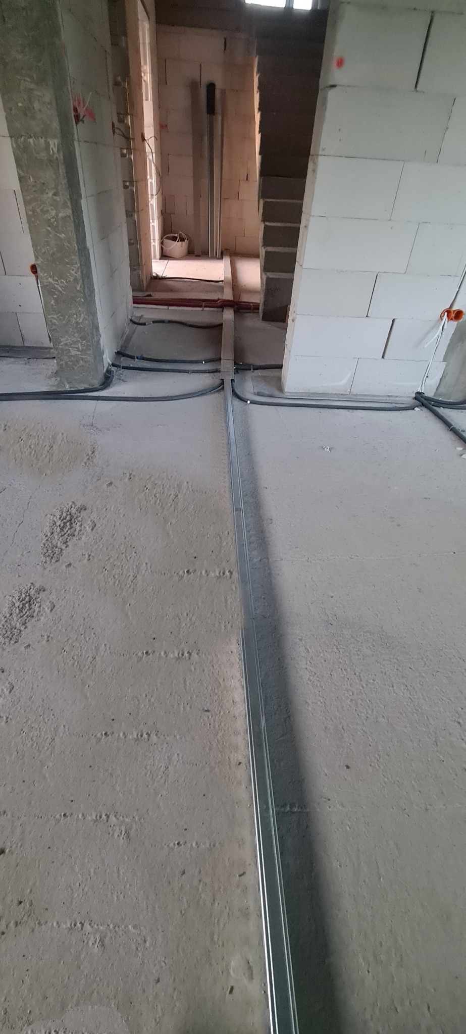 Instalacja elektryczna: Szare kable w peszlach prowadzone po surowym betonowym podłożu w trakcie budowy, z widoczną metalową listwą montażową.