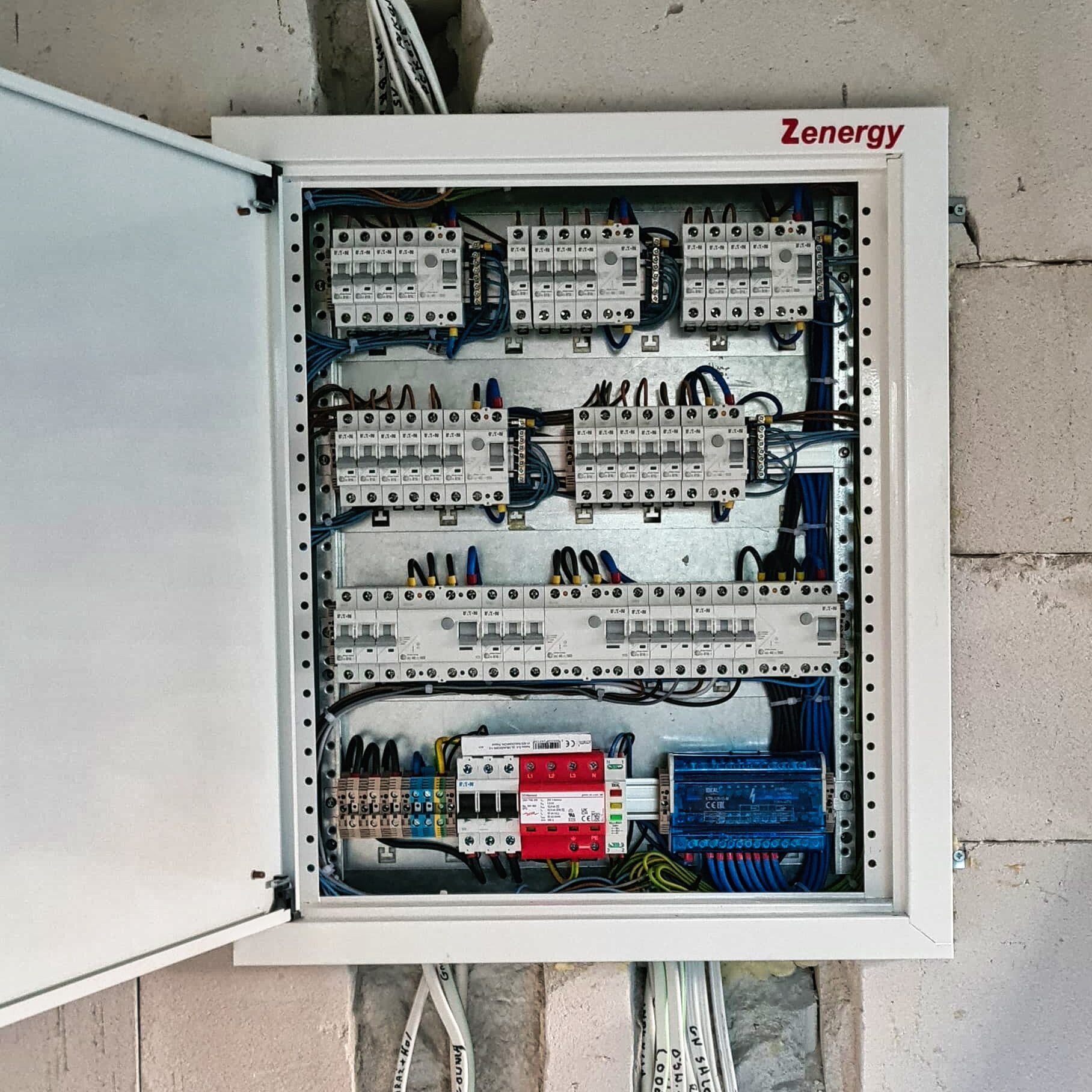Otwarta skrzynka bezpiecznikowa Zenergy z widocznymi bezpiecznikami, przewodami i elementami instalacji elektrycznej, zamontowana na surowej ścianie z betonu komórkowego.