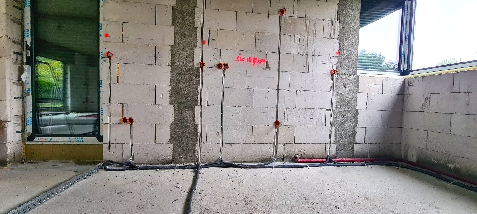 Wewnętrzne ściany budynku w trakcie budowy z widocznymi przewodami elektrycznymi w peszlach, puszkami instalacyjnymi w kolorze pomarańczowym i czerwonymi oznaczeniami na ścianie z betonu komórkowego.