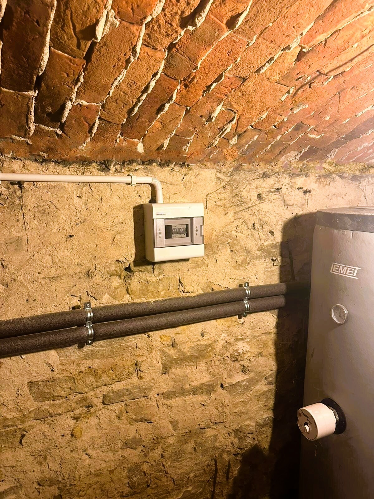 Skrzynka bezpiecznikowa z wyłącznikami zamontowana na starej, kamiennej ścianie w piwnicy z ceglanym sklepieniem łukowym, obok biegną rury centralnego ogrzewania z izolacją.