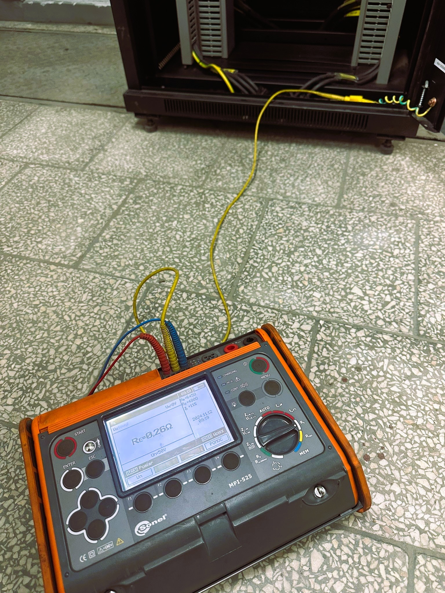 Miernik Sonel MPI-525 z podłączonymi przewodami pomiarowymi, wyświetlający wynik pomiaru rezystancji uziemienia 0,260Ω, ustawiony na posadzce z płytek, obok szafy rack z widocznym przewodem...