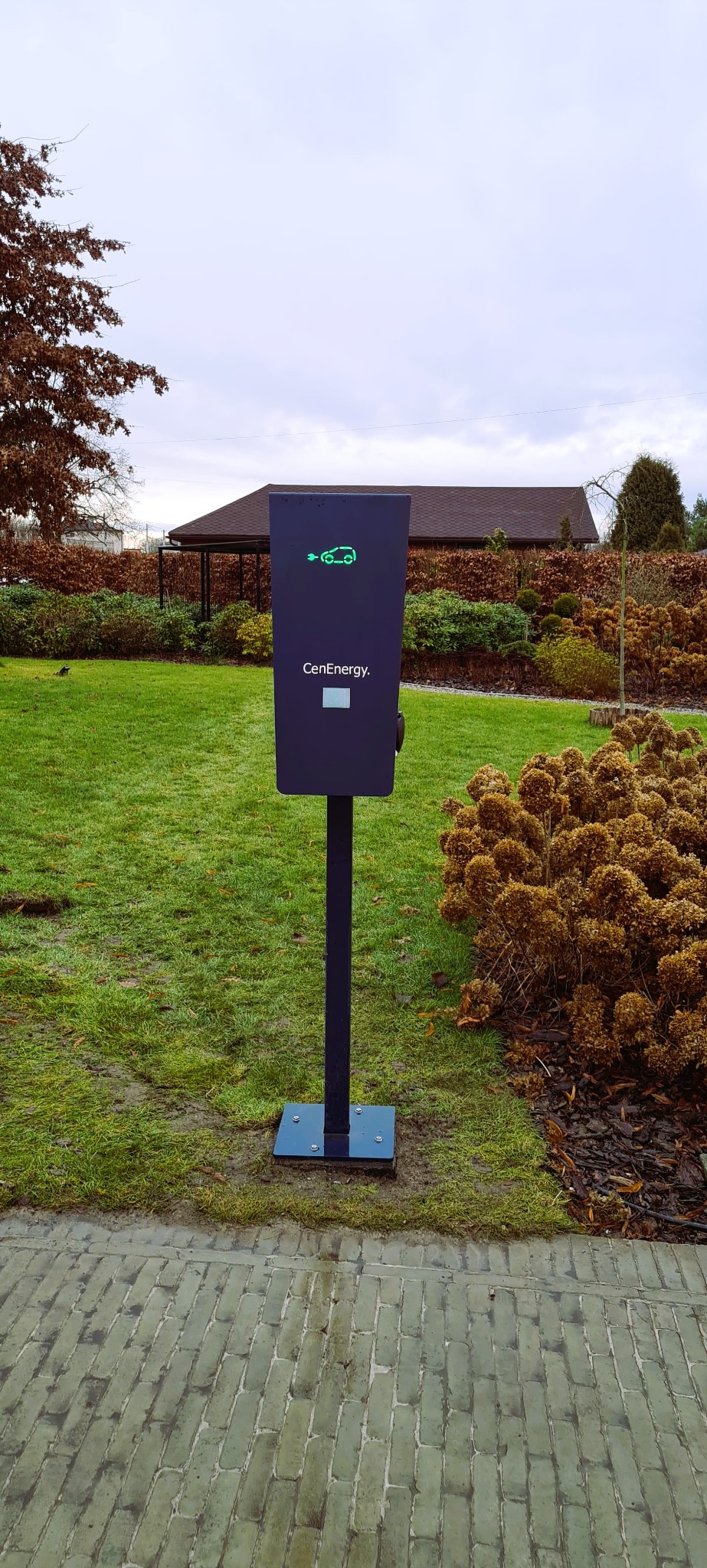 Stacja ładowania samochodów elektrycznych CenEnergy na tle zielonego trawnika i krzewów, z widocznym budynkiem w tle.