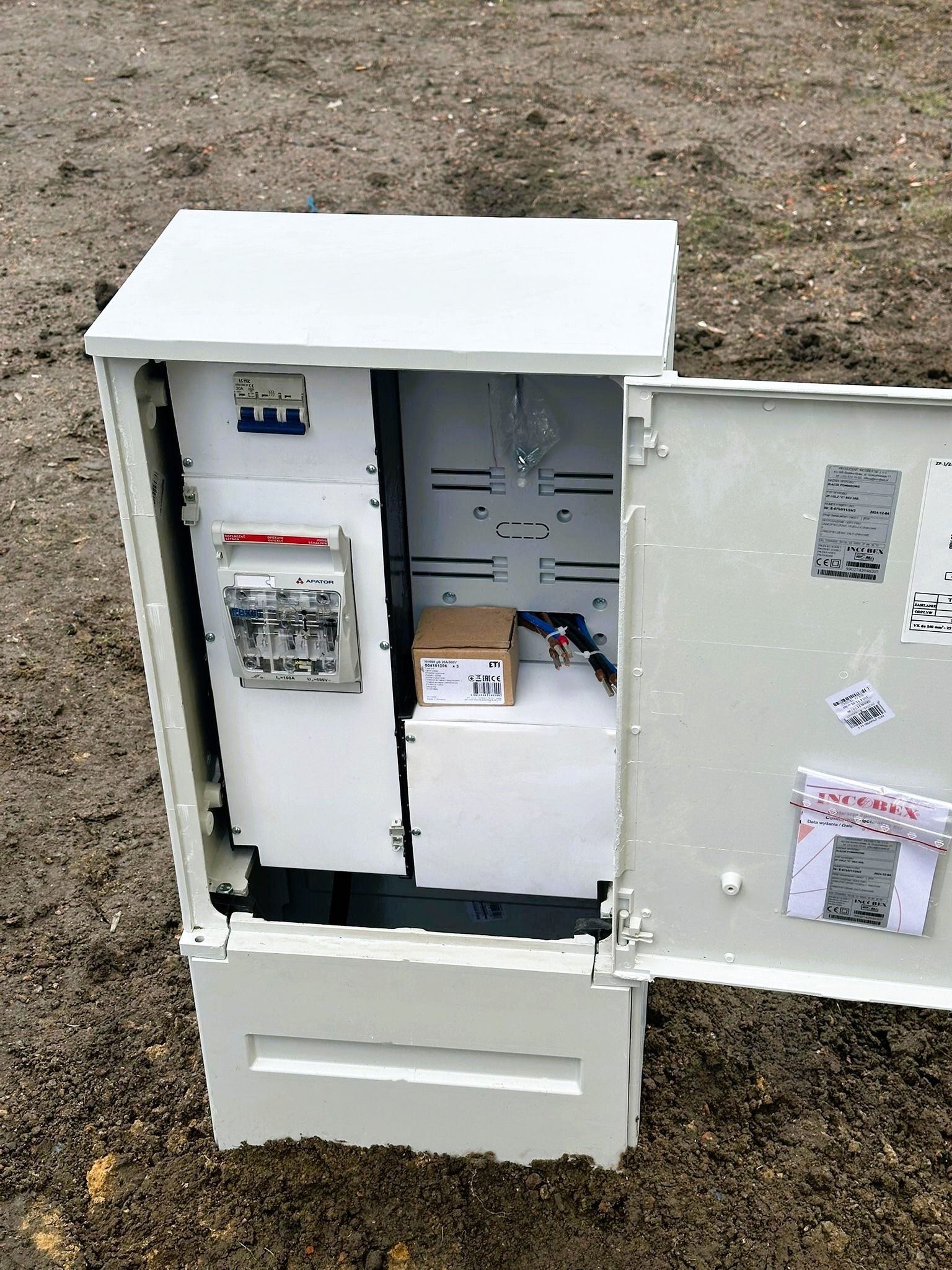 Otwarta, biała skrzynka elektryczna z widocznymi bezpiecznikami, licznikiem energii i przewodami na tle ziemi.