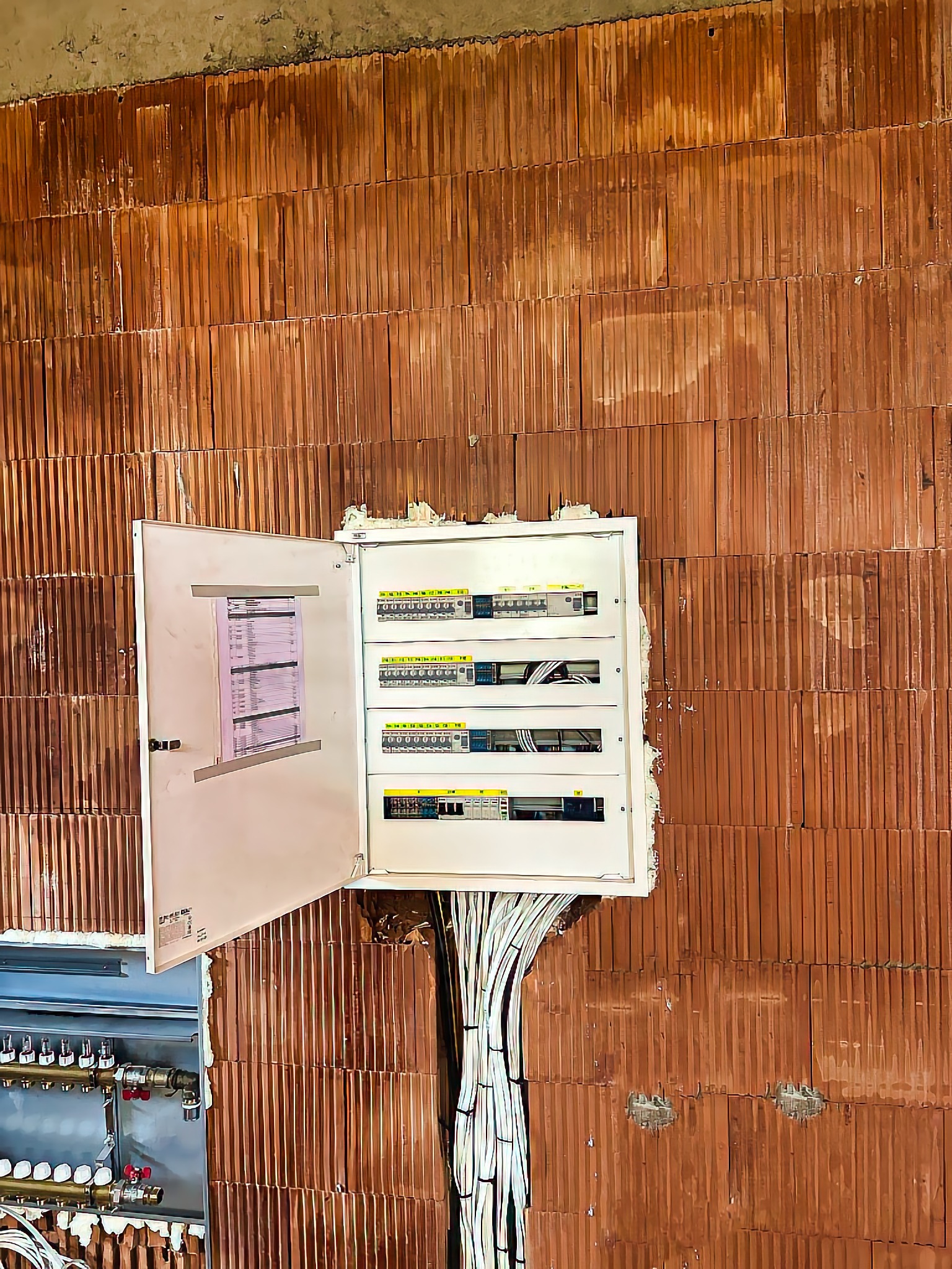 Otwarta skrzynka rozdzielcza z widocznymi bezpiecznikami i kablami, zamontowana na ścianie z czerwonej cegły dziurawki w surowym stanie.