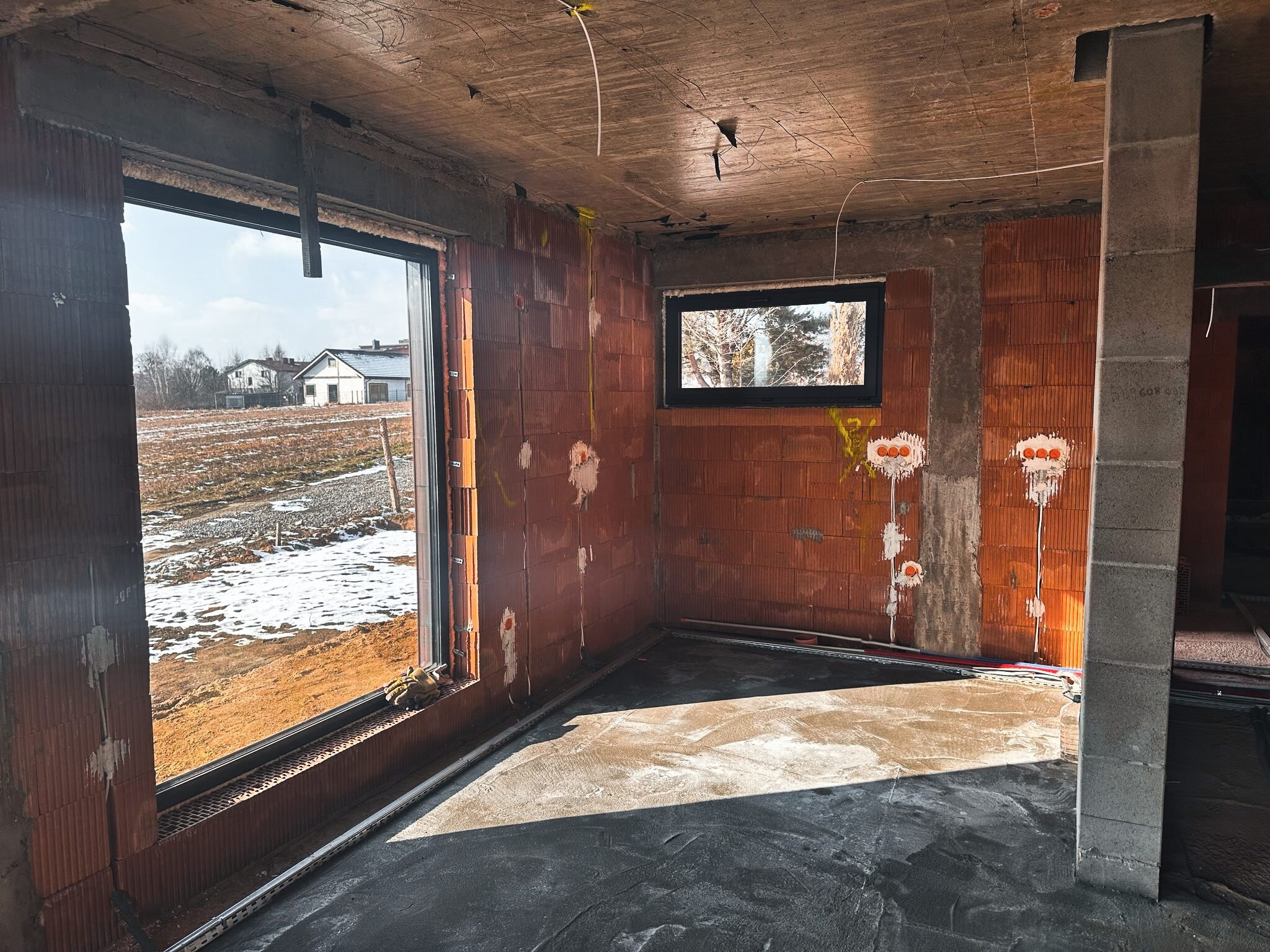 Surowy stan wnętrza budynku w trakcie budowy: widoczne ceglane ściany z bruzdami na przewody, betonowa podłoga, okna z widokiem na krajobraz zimowy, wystające kable z sufitu.