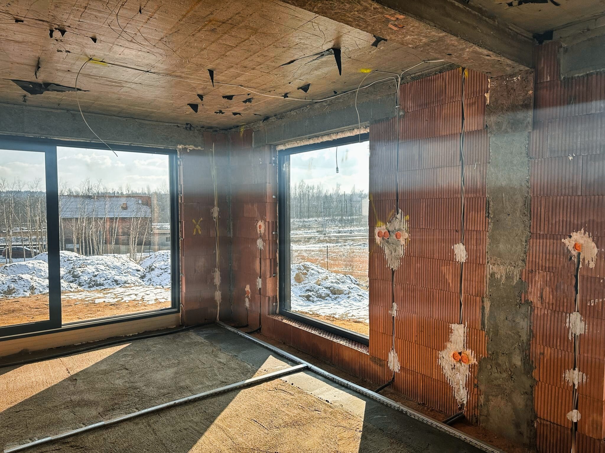 Wnętrze surowego domu w trakcie instalacji elektrycznej: widoczne przewody, puszki instalacyjne w ścianach z pustaków ceramicznych, częściowo wylana podłoga, okna z widokiem na zimowy krajobraz.