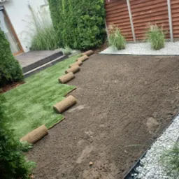 Rozwinięta trawa z rolki na przygotowanym podłożu, widoczna granica z białych kamieni i drewniany płot w tle, fragment domu z drewnianym tarasem po lewej stronie.