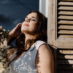 Maciej Wadas Fotografia - Portret kobiety w białej sukience, opierającej się o drewniane okiennice z zamkniętymi oczami w promieniach słońca, w tle dekoracja z suszonych traw.