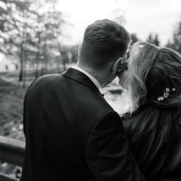 Maciej Wadas Fotografia - Czarno-białe zdjęcie pary całującej się na tle drzew i wody. Mężczyzna w garniturze, kobieta w skórzanej kurtce z ozdobą we włosach.