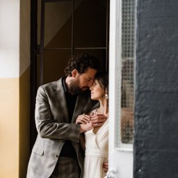 Maciej Wadas Fotografia - Para w objęciach przy oknie: mężczyzna w garniturze w kratę obejmuje kobietę w białej sukience, oboje z zamkniętymi oczami, w tle okno z kratami.