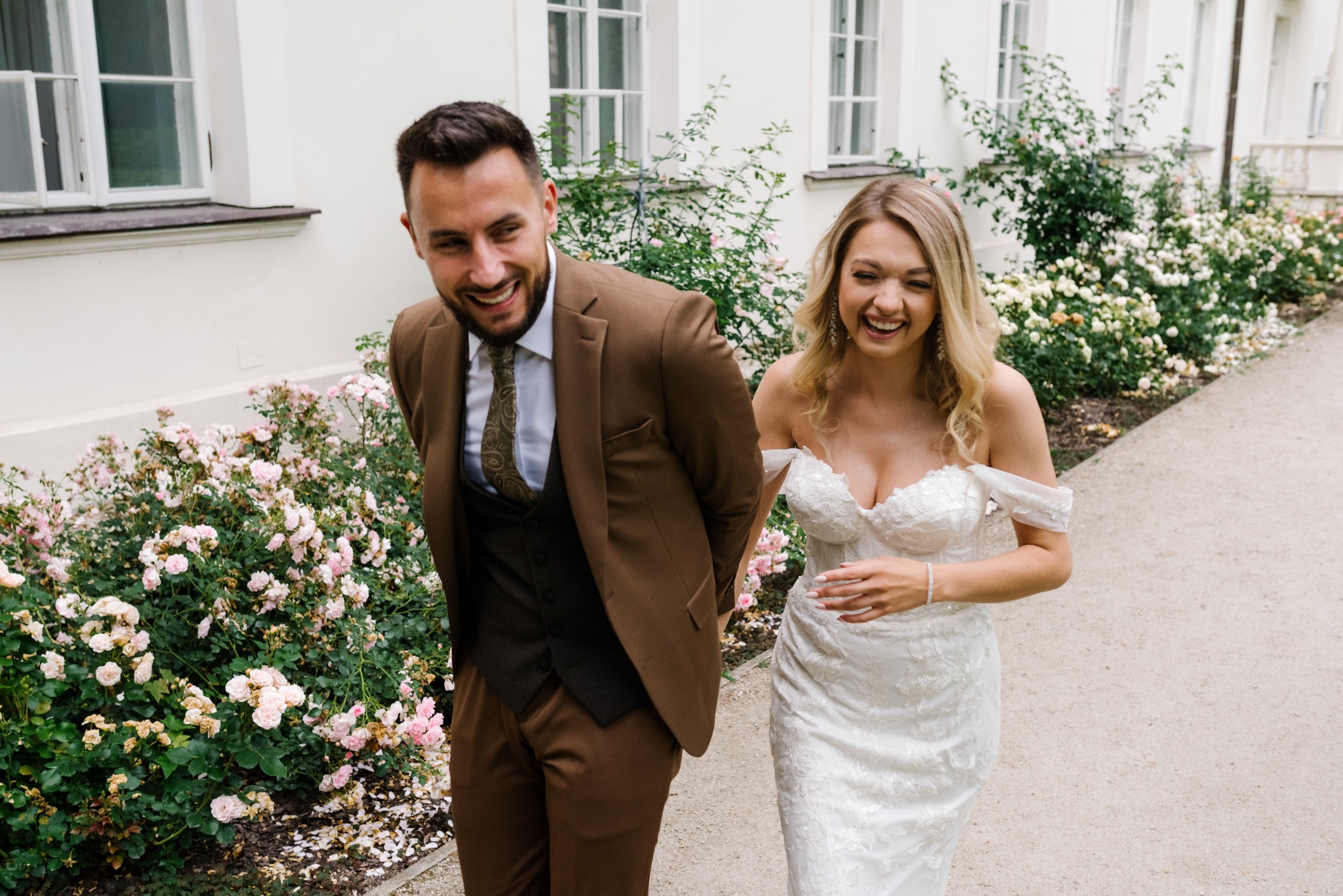 Uśmiechnięta para młoda w strojach ślubnych spaceruje po parku, biała suknia z koronkowymi zdobieniami, brązowy garnitur, jasne tło budynku i kwitnące krzewy róż.