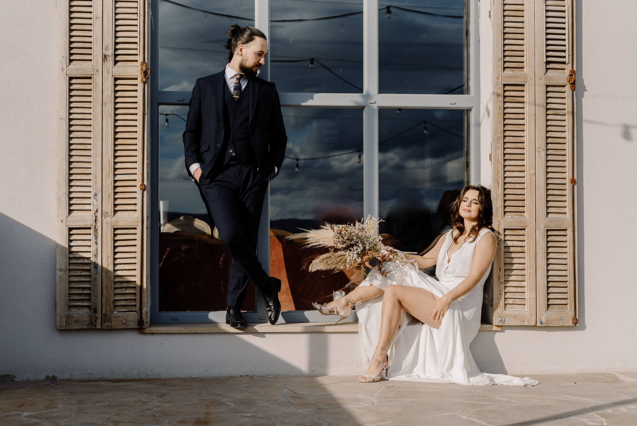 Panna młoda w białej sukni siedzi na parapecie okna z drewnianymi okiennicami, trzyma bukiet z suszonych kwiatów, obok stoi pan młody w granatowym garniturze.