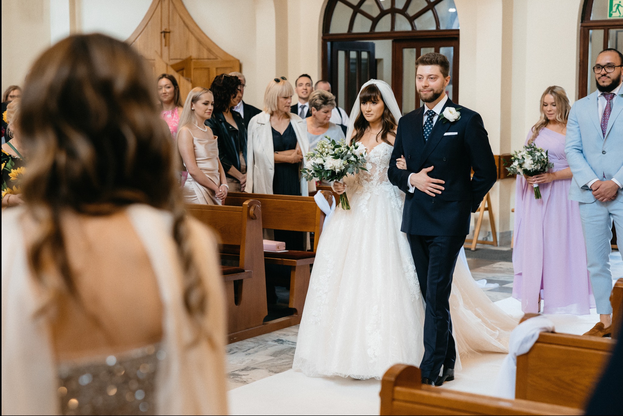 Panna młoda w białej sukni z welonem i bukietem prowadzona do ołtarza przez pana młodego w granatowym garniturze, w tle goście weselni w kościele.
