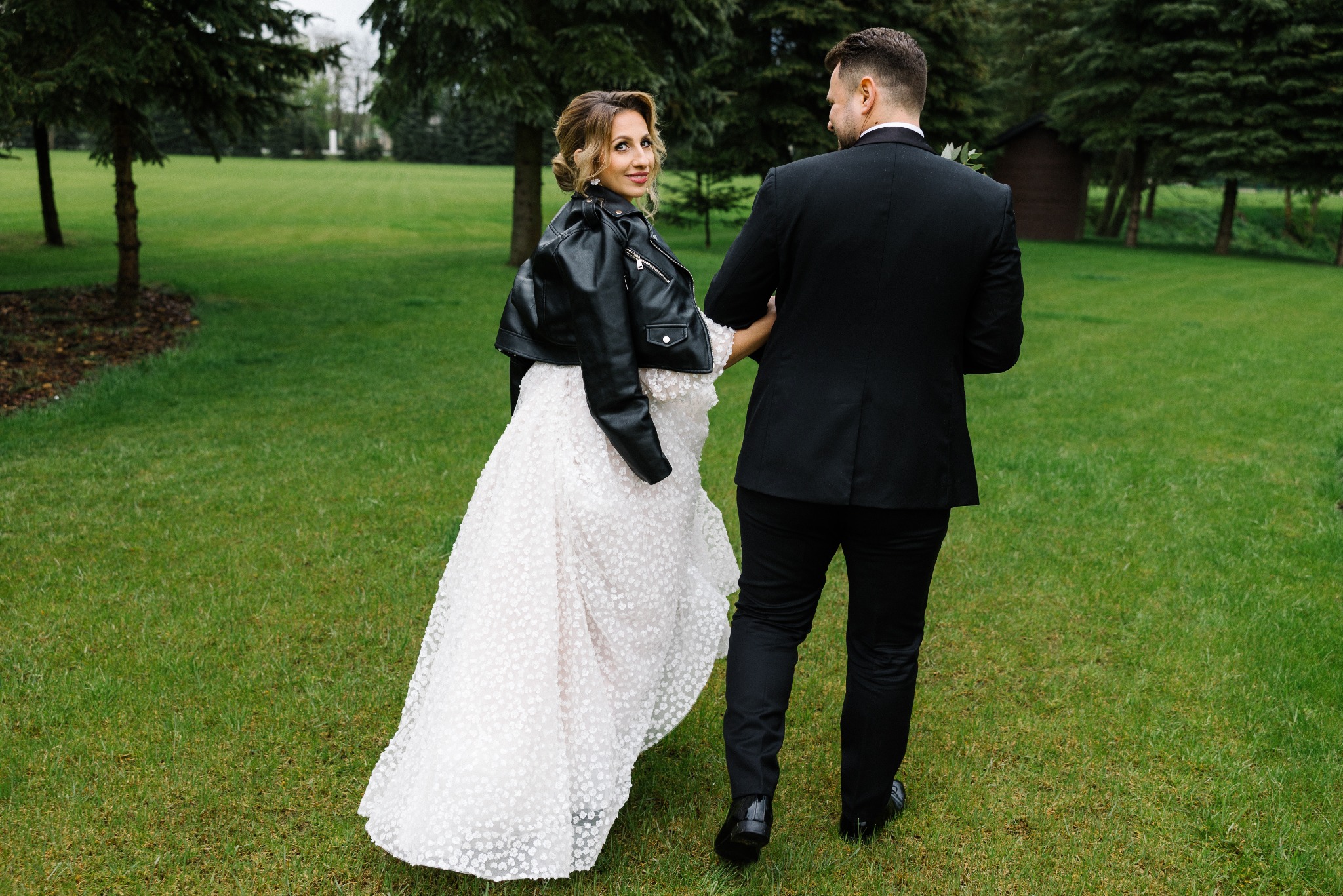 Panna młoda w białej sukni z koronkowymi aplikacjami i czarnej skórzanej kurtce, odwraca się spoglądając przez ramię na idącego obok pana młodego w czarnym garniturze, na tle zielonego trawnika...
