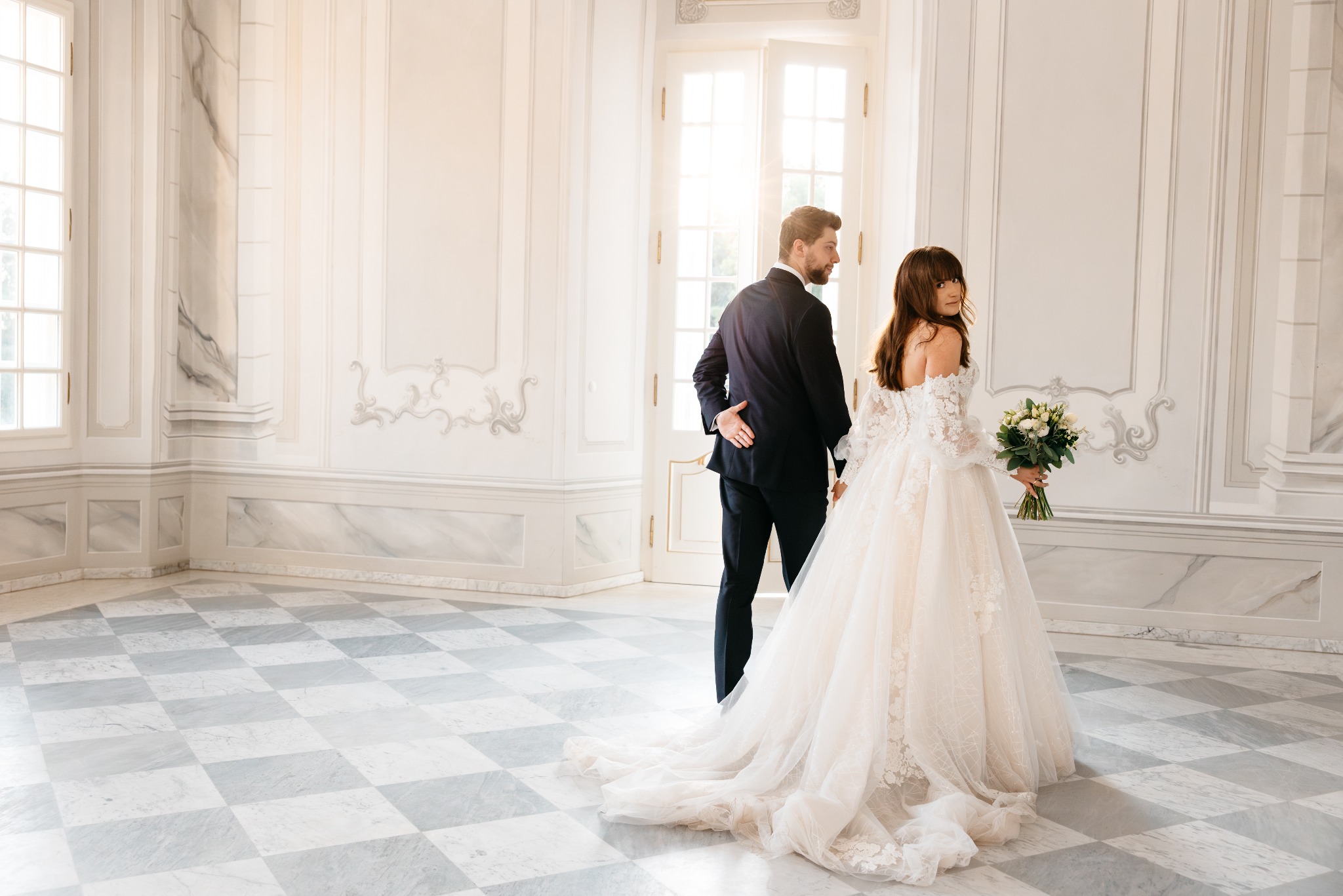 Para młoda w eleganckim, jasnym wnętrzu z marmurową podłogą w szachownicę; panna młoda w sukni z koronkowymi rękawami i bukietem odwraca się, patrząc przez ramię.