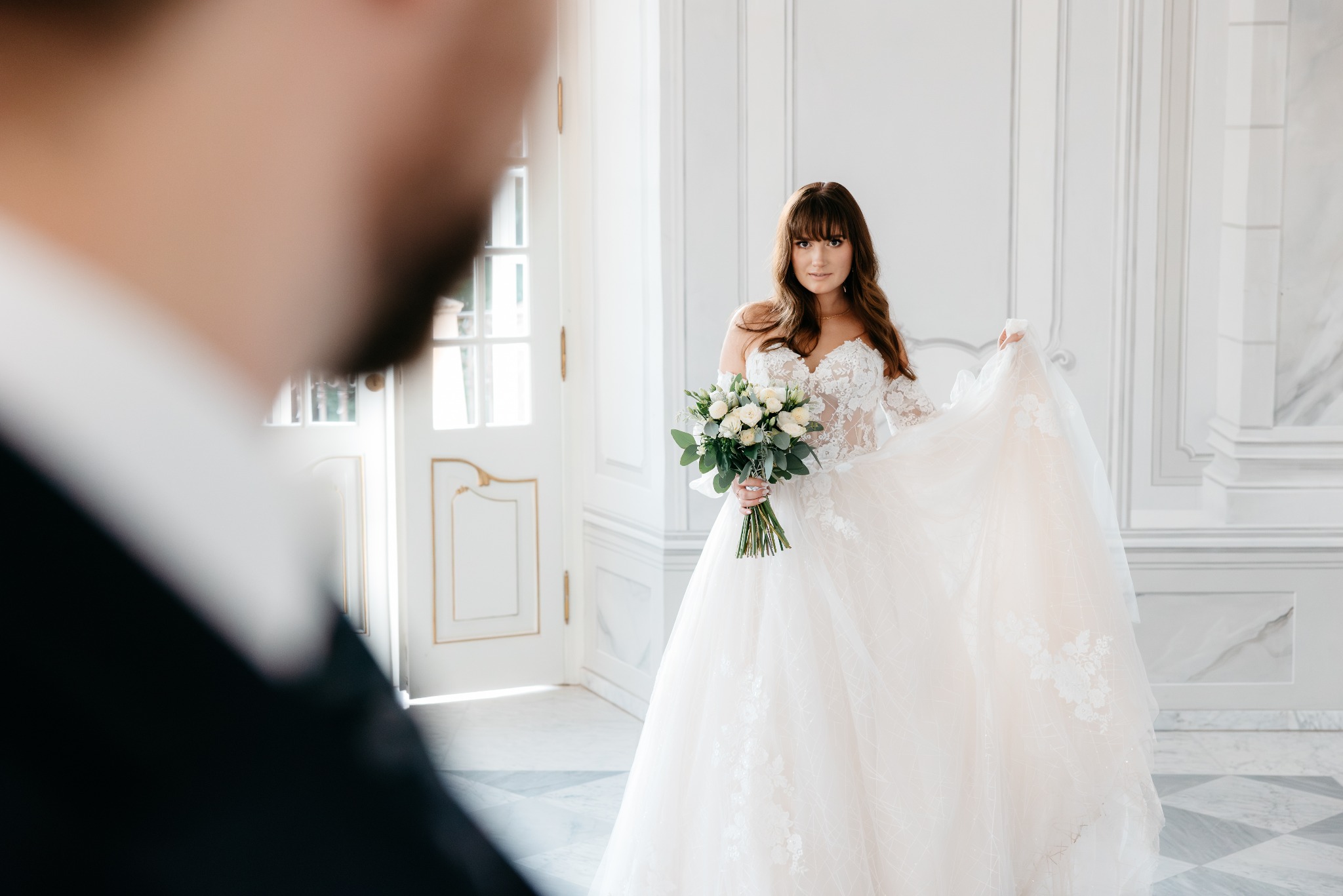 Panna młoda w koronkowej sukni balowej, trzymająca bukiet białych róż i zielonych liści, pozuje w jasnym wnętrzu z marmurową podłogą i białymi drzwiami z dekoracyjnymi złotymi elementami, widziana...