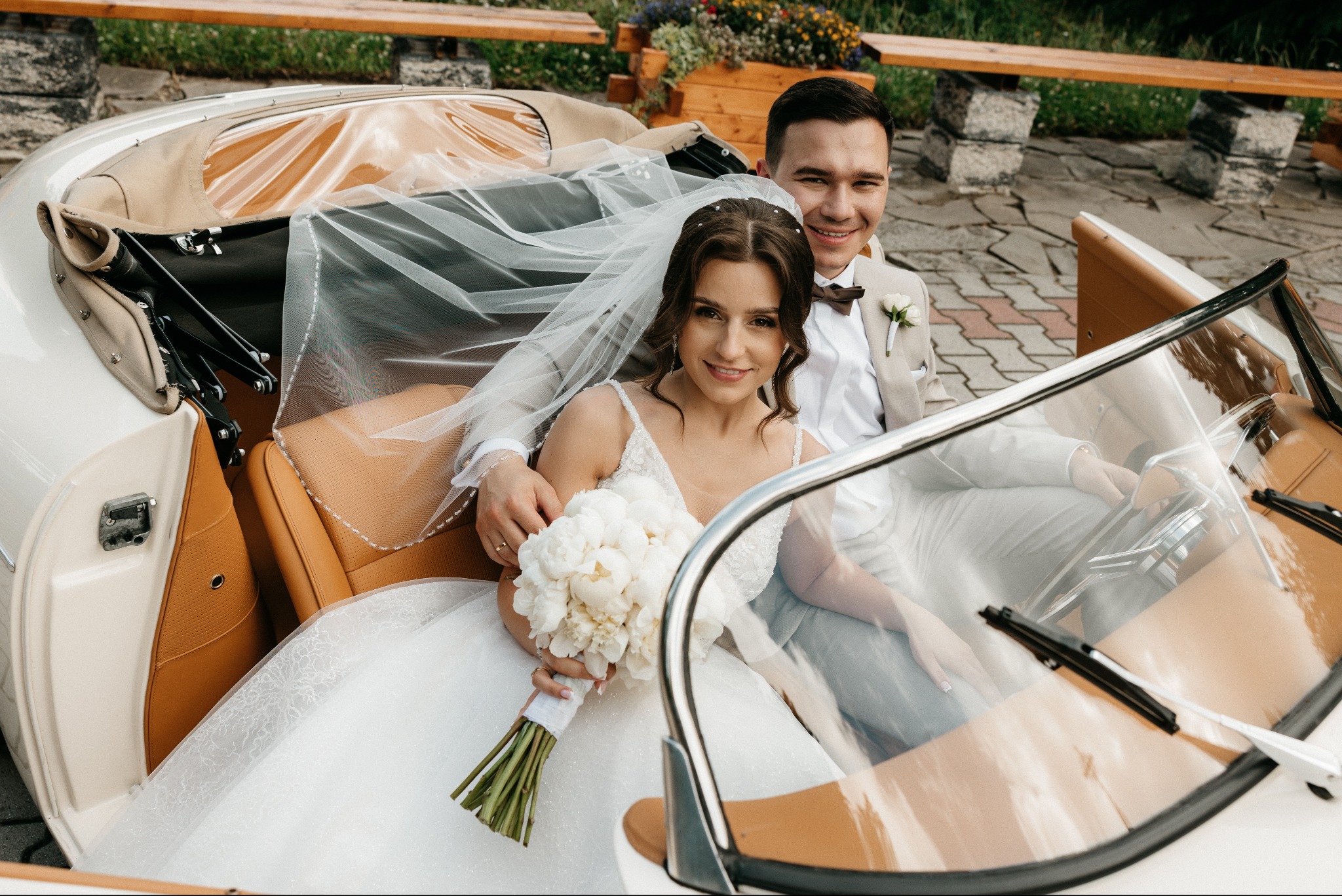 Uśmiechnięta para młoda siedzi w białym kabriolecie z brązową skórzaną tapicerką, panna młoda trzyma bukiet białych piwonii, a pan młody ma beżowy garnitur i brązową muchę.