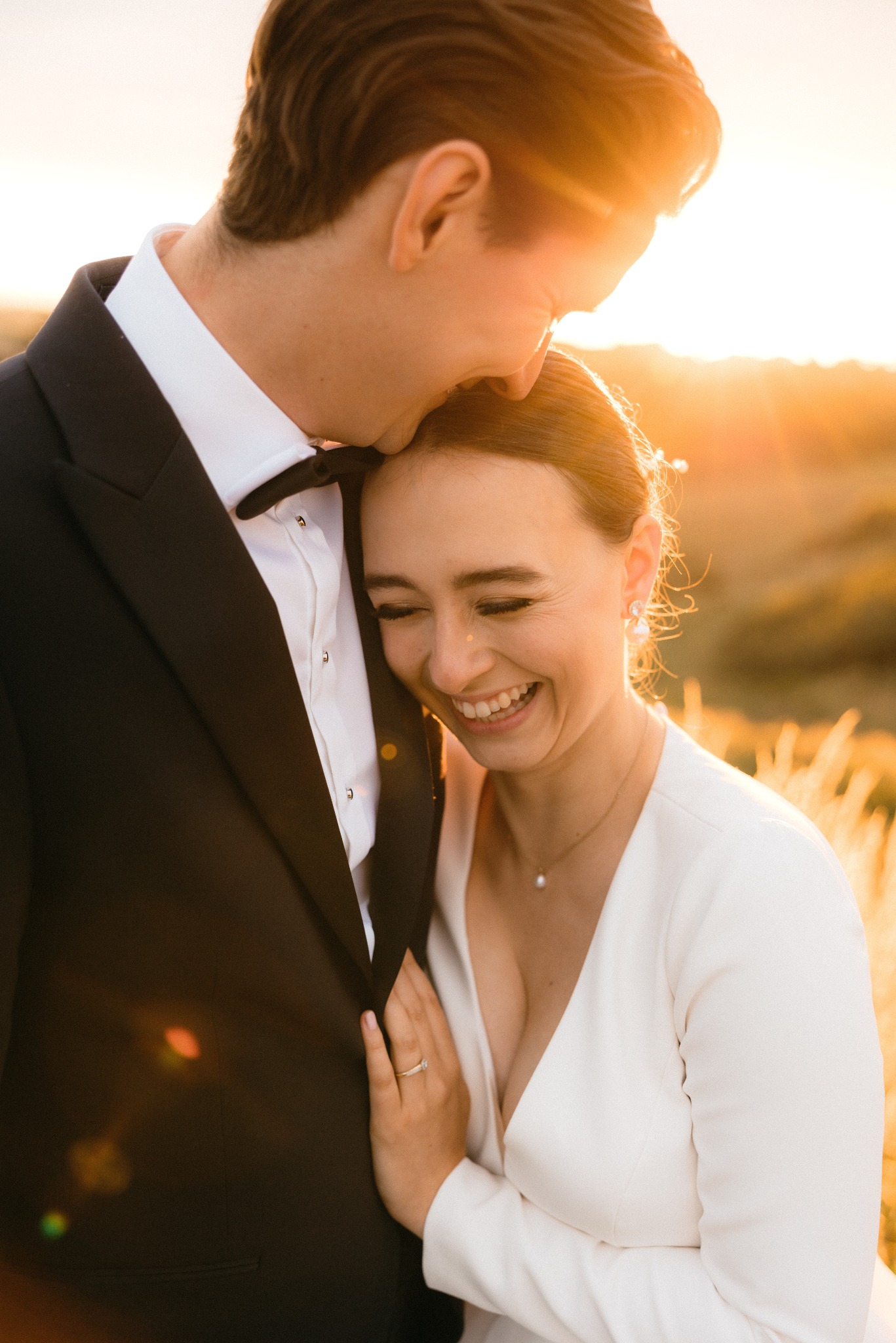 Uśmiechnięta panna młoda opiera głowę na ramieniu pana młodego w czarnym smokingu z muszką, złote światło zachodzącego słońca w tle, pierścionek zaręczynowy na palcu panny młodej.