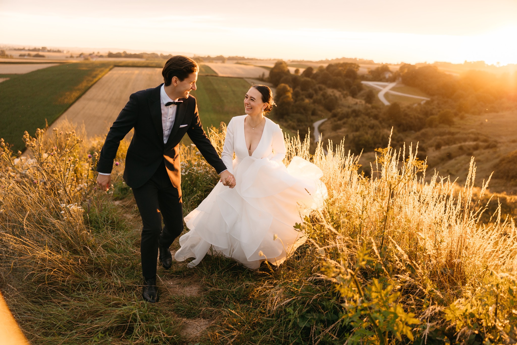 Uśmiechnięta para młoda, trzymając się za ręce, biegnie po wzgórzu w promieniach zachodzącego słońca; w tle pola uprawne i las.