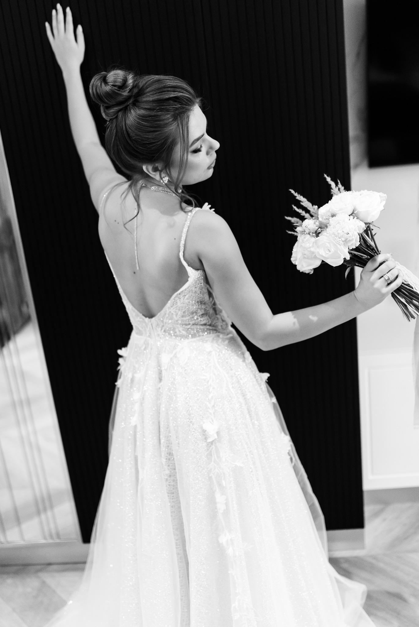 Panna młoda w koronkowej sukni z odkrytymi plecami, trzyma bukiet białych róż, pozuje na czarno-białym zdjęciu z uniesioną ręką i upiętymi włosami.