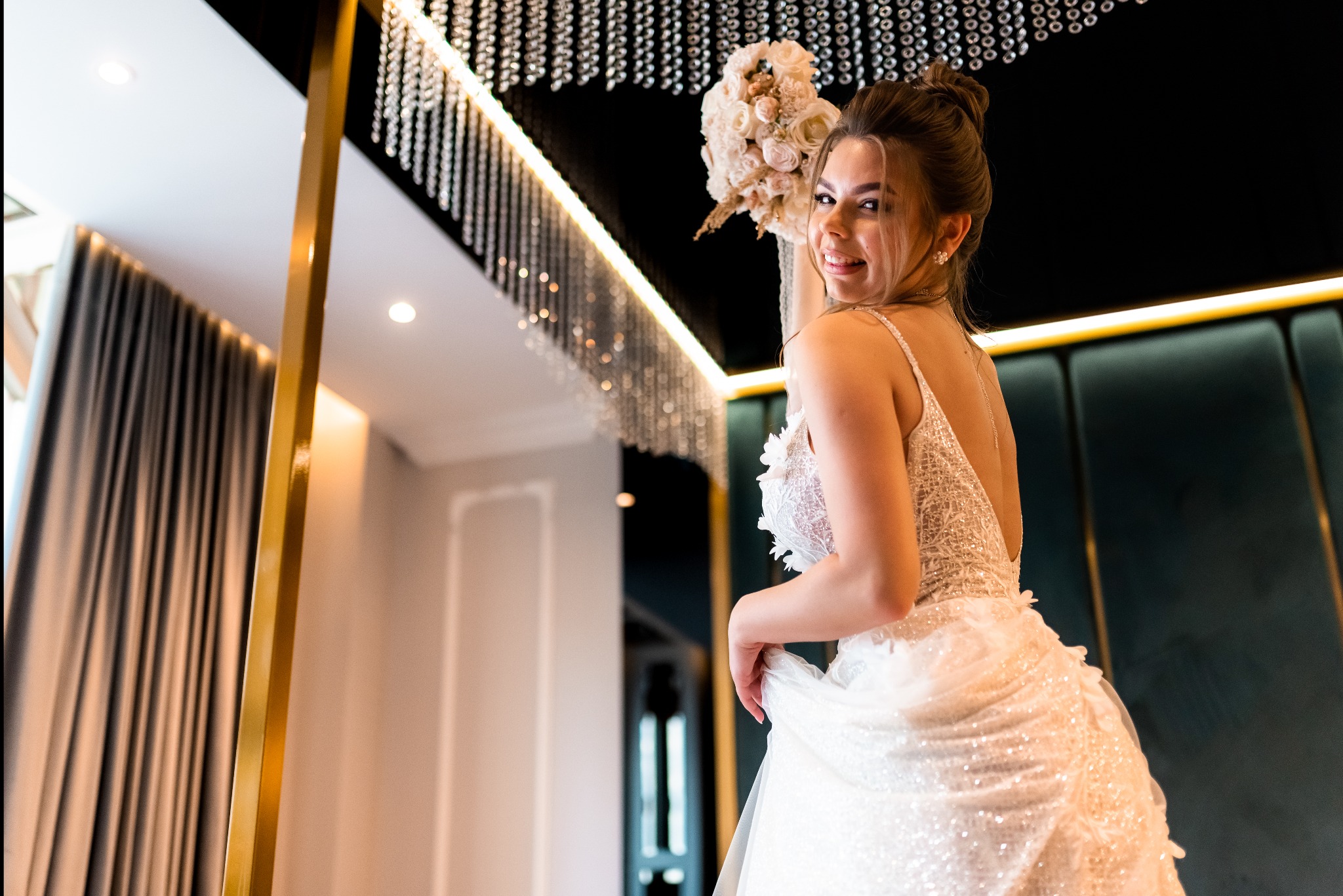 Uśmiechnięta panna młoda w błyszczącej sukni, trzymająca bukiet ślubny, spogląda przez ramię na tle kryształowego żyrandola i ciemnozielonej ściany.