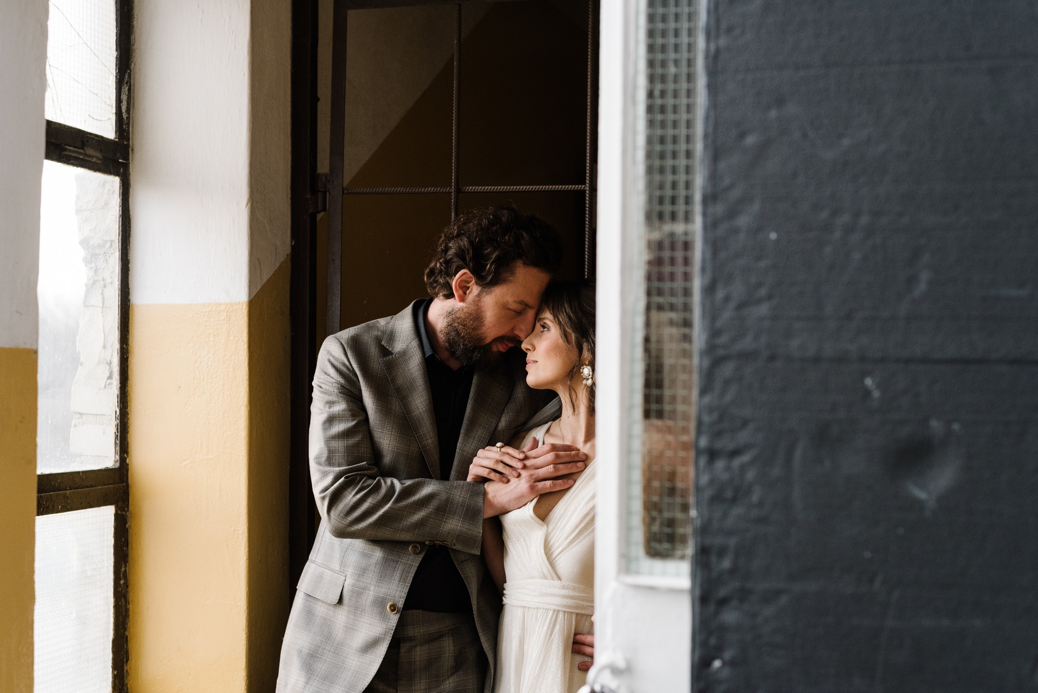 Para w objęciach przy oknie: mężczyzna w garniturze w kratę obejmuje kobietę w białej sukience, oboje z zamkniętymi oczami, w tle okno z kratami.