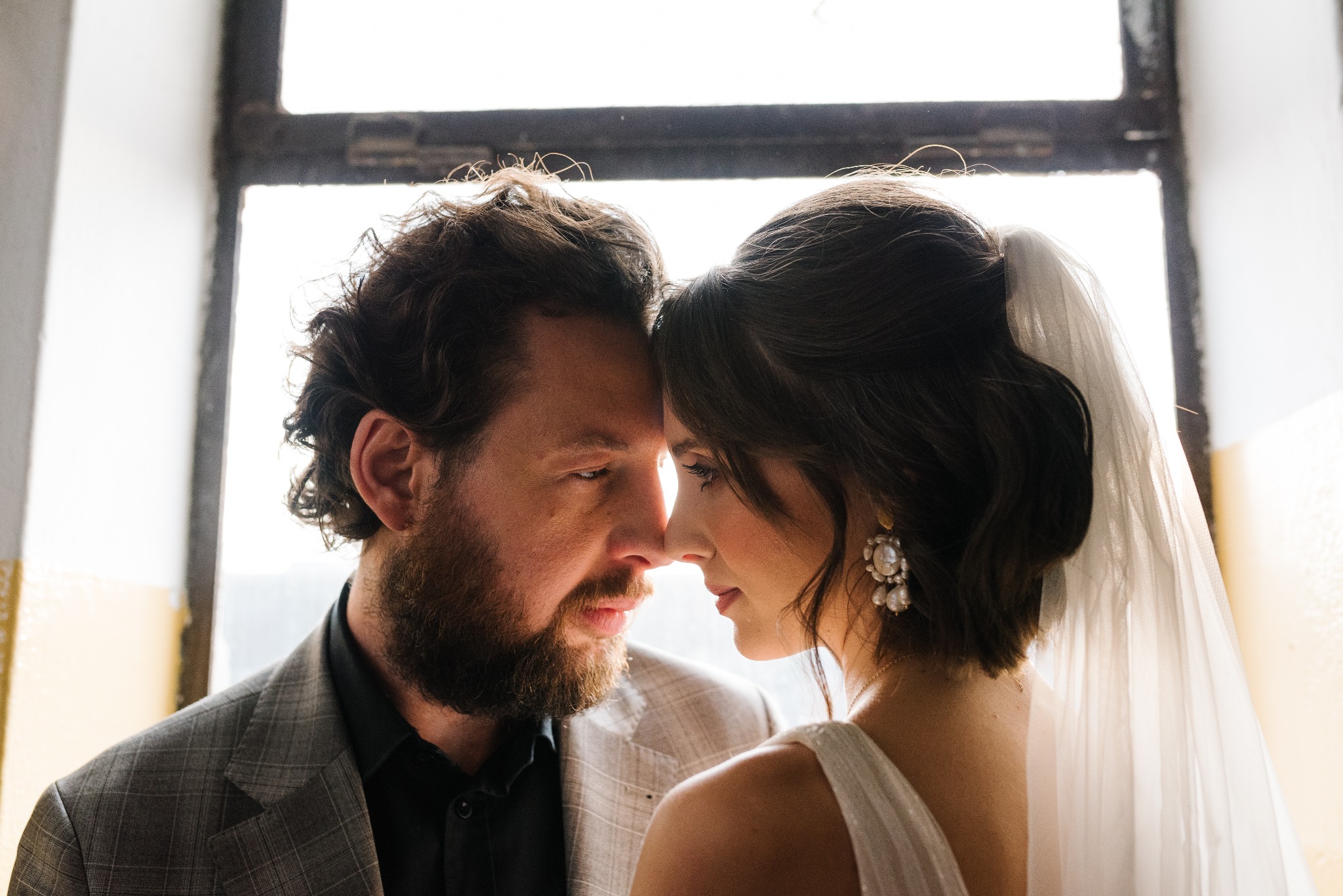 Zbliżenie na twarze pary: brodaty mężczyzna w szarej marynarce i kobieta w białej sukni ślubnej z welonem i perłowymi kolczykami, dotykający się nosami na tle okna z jasnym światłem.