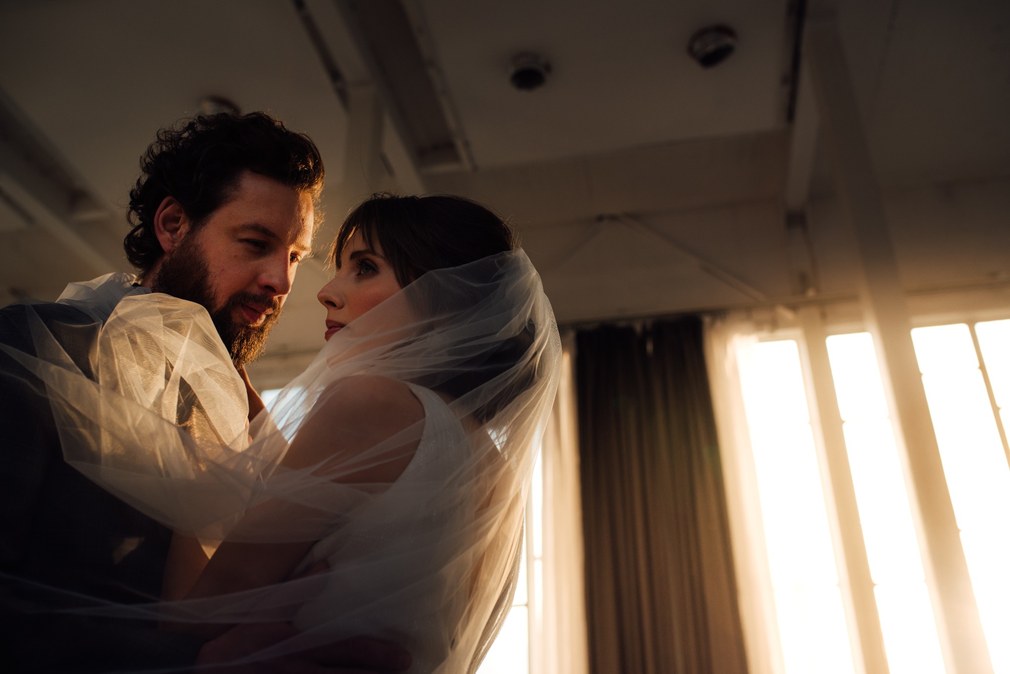 Para młoda w objęciach, panna młoda okryta welonem, spojrzenia skierowane ku sobie, ciepłe światło z okna w tle.