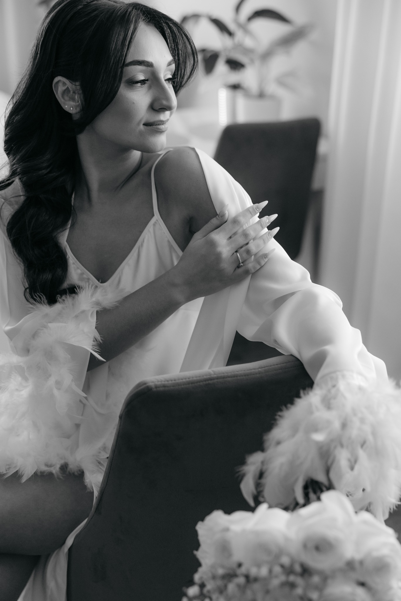 Czarno-białe zdjęcie kobiety w szlafroku z piórami, z pierścionkiem zaręczynowym na palcu, siedzącej na krześle, spoglądającej w bok. Romantyczna atmosfera.