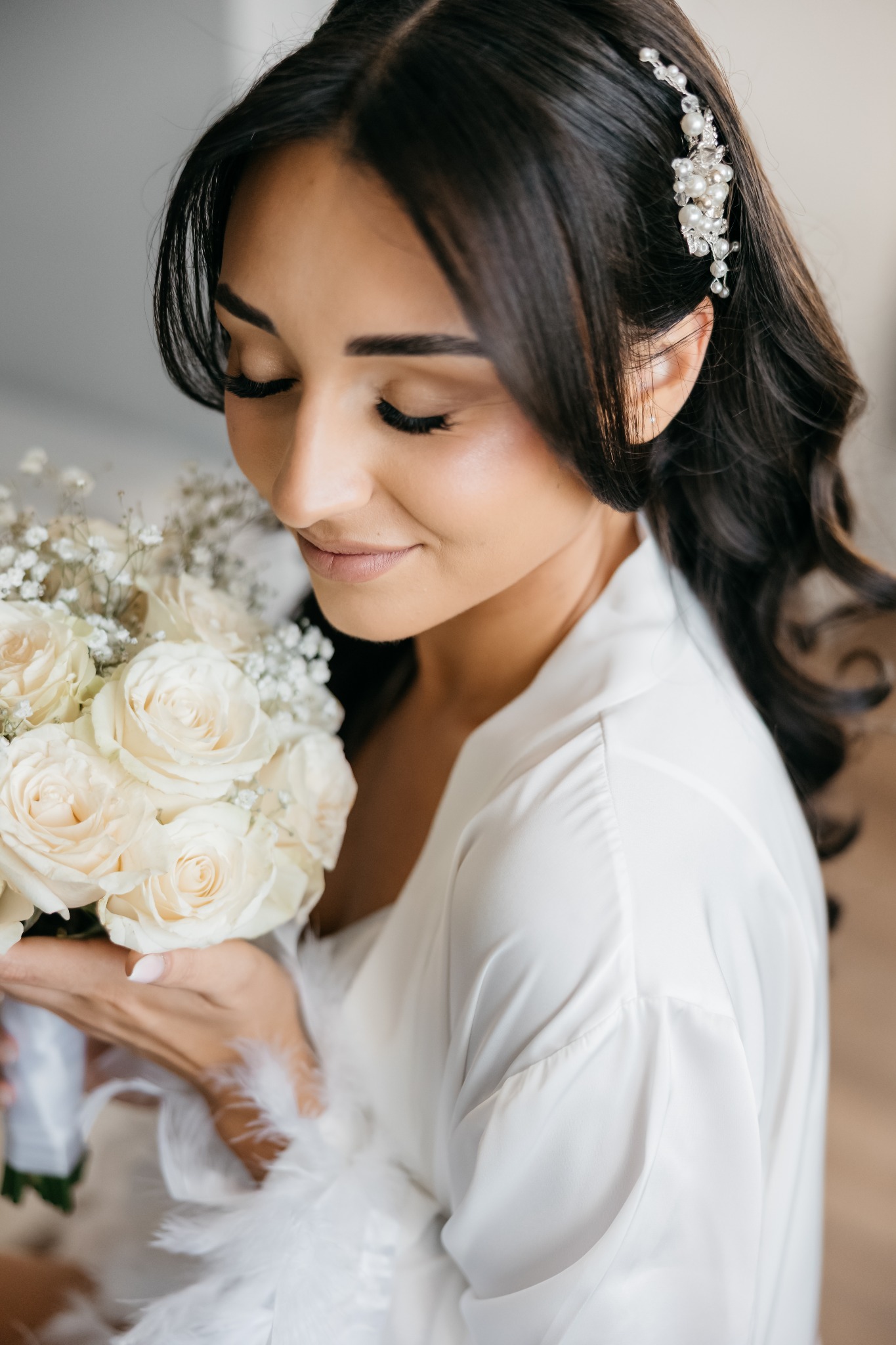 Panna młoda w białym szlafroku, z bukietem białych róż i gipsówki, ozdobą z pereł we włosach, spogląda w dół z delikatnym uśmiechem.