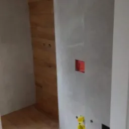 Fragment łazienki w trakcie wykańczania: szare płytki na ścianach z widocznymi otworami na instalacje, drewnopodobne płytki na fragmencie ściany i podłodze, widoczne żółte elementy instalacji wodnej.