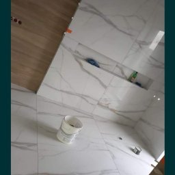 Tomasz Tomasz - Świeżo ułożone, marmurowe płytki w łazience z wnękami, widoczne narzędzia i wiadro, częściowo wykończona podłoga z paneli.