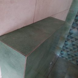 Tomasz Tomasz - Zielona, ceramiczna zabudowa w rogu pomieszczenia, z widocznymi fugami i fragmentem wzorzystej podłogi w tle.