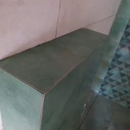 Zielona, ceramiczna zabudowa w rogu pomieszczenia, z widocznymi fugami i fragmentem wzorzystej podłogi w tle.