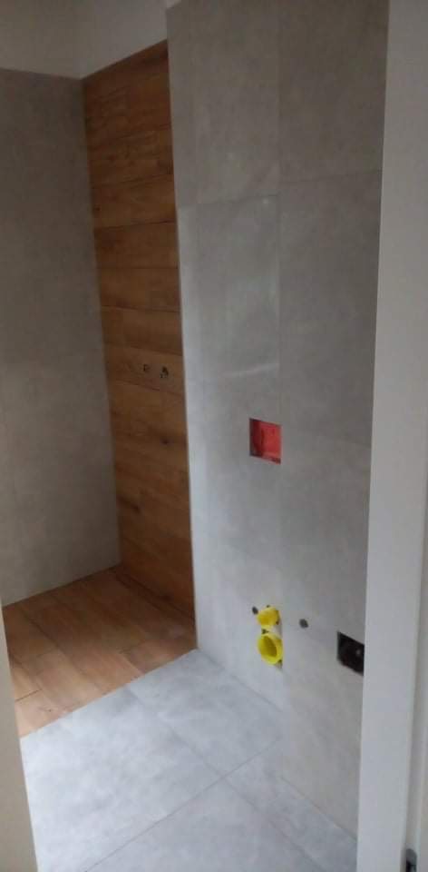 Fragment łazienki w trakcie wykańczania: szare płytki na ścianach z widocznymi otworami na instalacje, drewnopodobne płytki na fragmencie ściany i podłodze, widoczne żółte elementy instalacji wodnej.