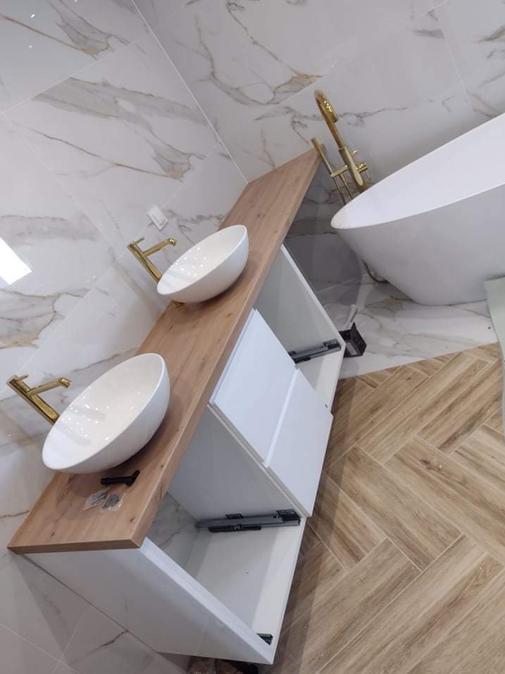 Nowoczesna łazienka z dwoma białymi umywalkami nablatowymi o owalnym kształcie, zamontowanymi na drewnianym blacie z białymi szafkami, złota armatura, biała wanna wolnostojąca i marmurowe płytki...