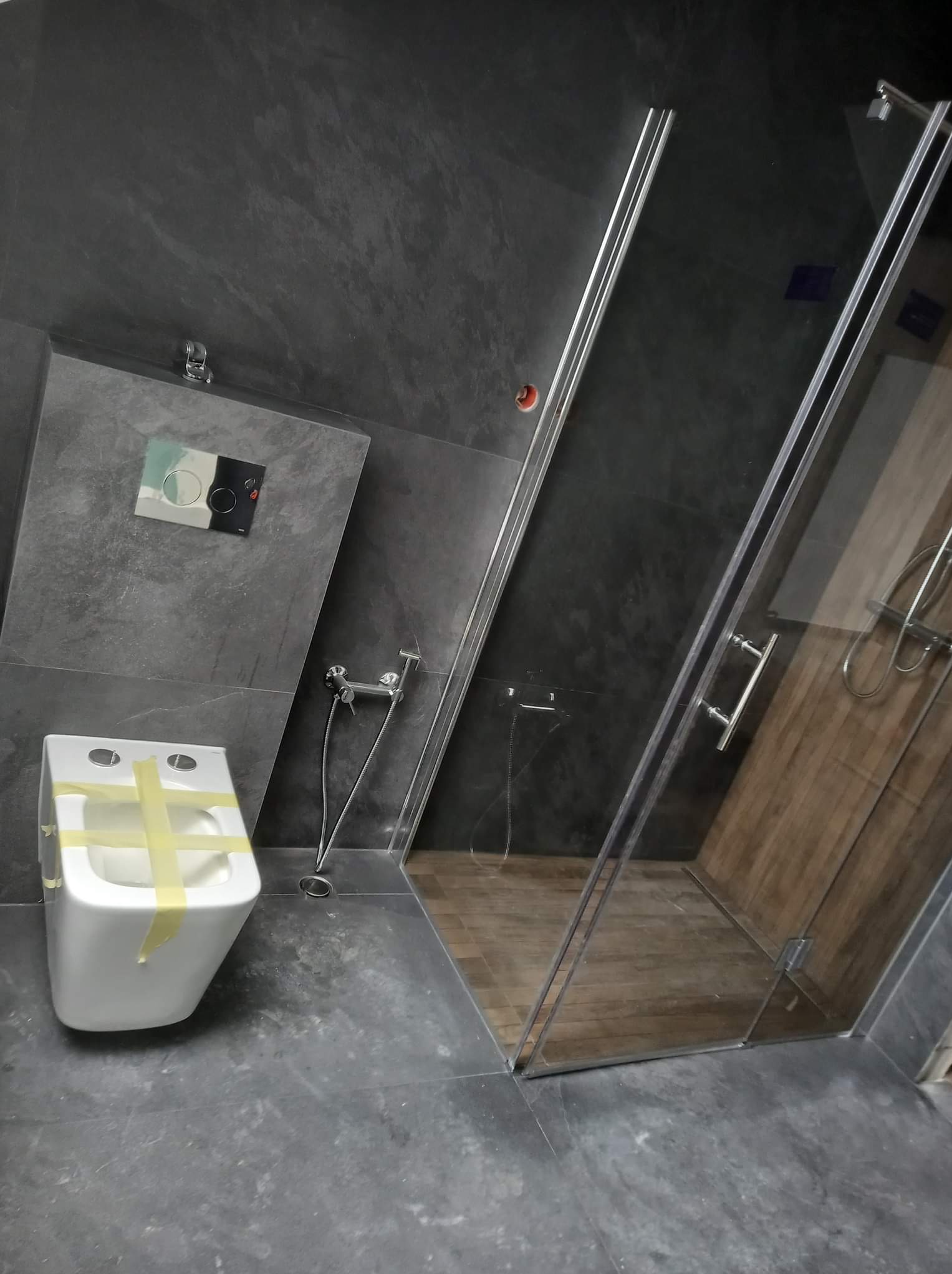 Nowoczesna łazienka w trakcie wykańczania: szare płytki na ścianach i podłodze, biała podwieszana toaleta zabezpieczona taśmą, szklana kabina prysznicowa z drewnopodobnym wykończeniem wewnątrz.