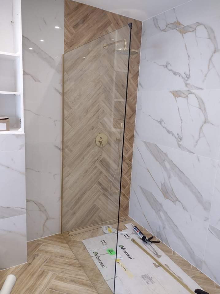 Nowoczesna kabina prysznicowa ze złotą armaturą i płytkami imitującymi drewno ułożonymi w jodełkę, kontrastująca z marmurowymi płytkami na ścianach, widoczne narzędzia i materiały montażowe...