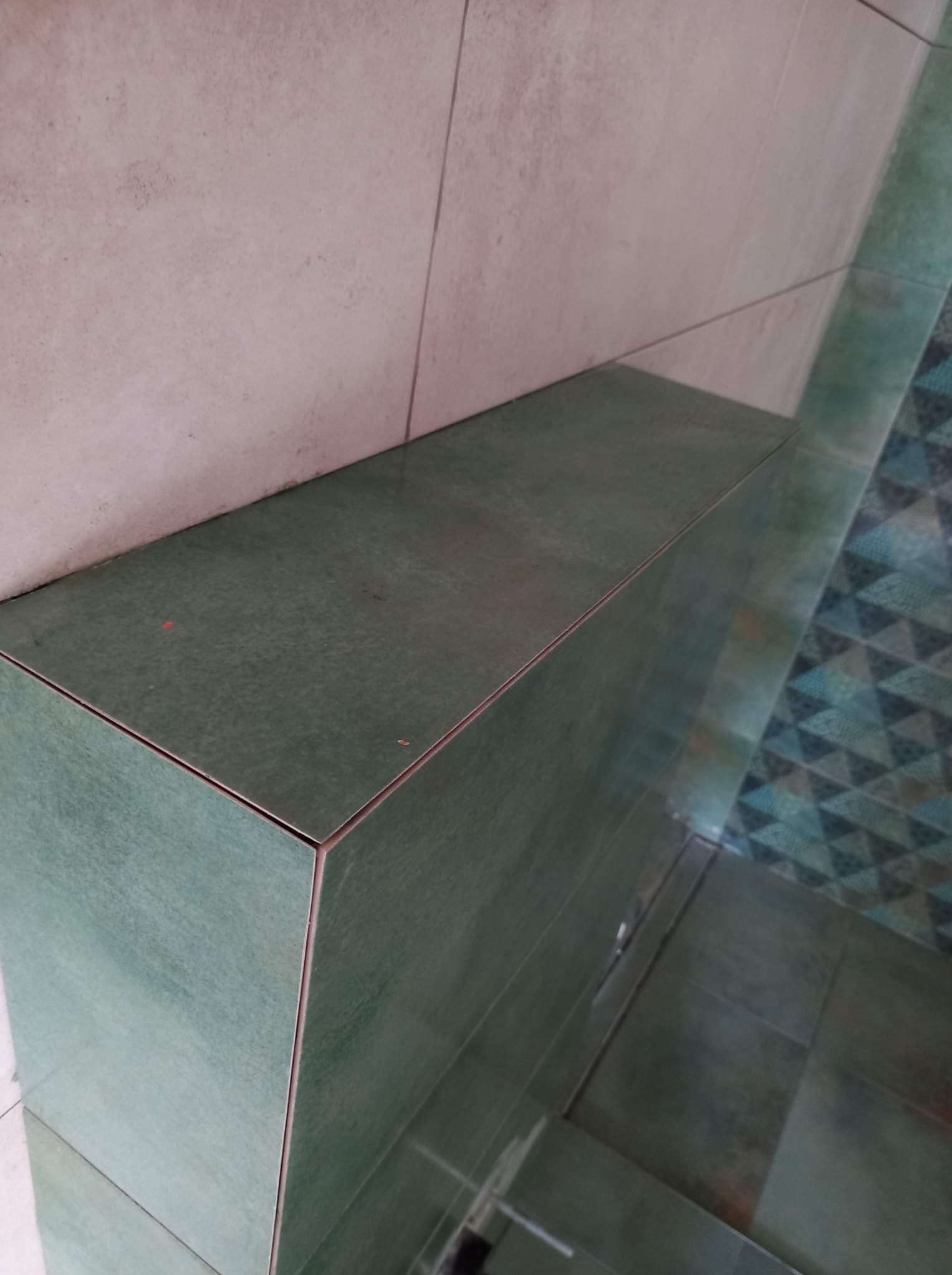 Zielona, ceramiczna zabudowa w rogu pomieszczenia, z widocznymi fugami i fragmentem wzorzystej podłogi w tle.