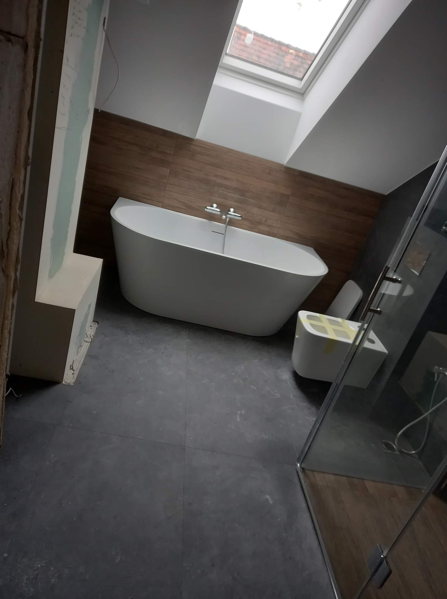 Wnętrze łazienki w trakcie prac wykończeniowych: wolnostojąca wanna, podwieszana toaleta z zabezpieczeniem, częściowo wykończona ściana z widoczną konstrukcją i płytą gipsową, oraz kabina...